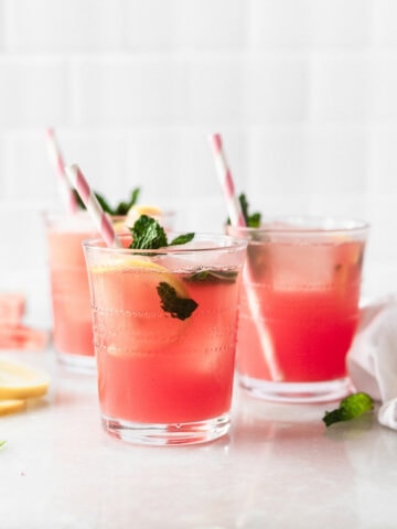 three glasses of watermelon mint lemonade with pink striped straws surrounded by lemon slices and mint leaves.
