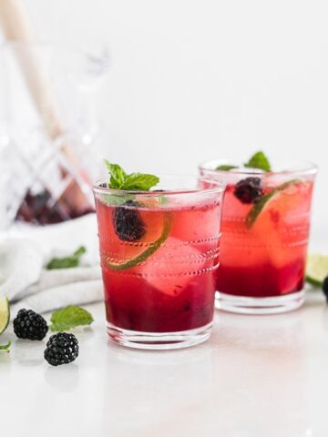 two blackberry bourbon smash cocktails in glasses with lime slices and blackberries with a glass shaker in the background.