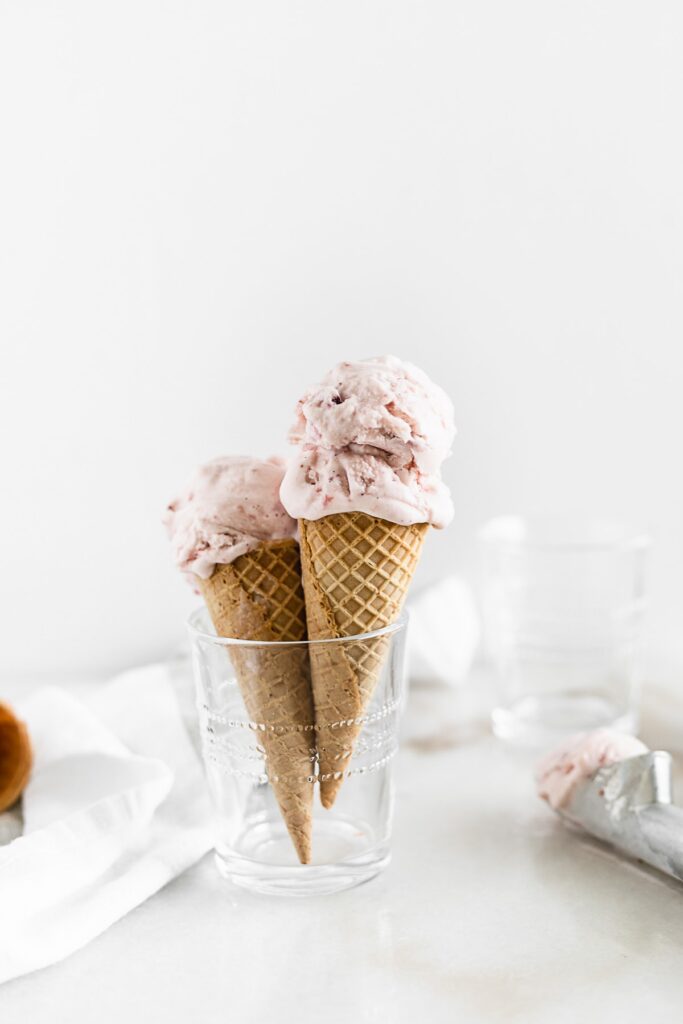 two roasted strawberry ice cream cones in a glass in front of a white background with an ice cream scoop beside them.