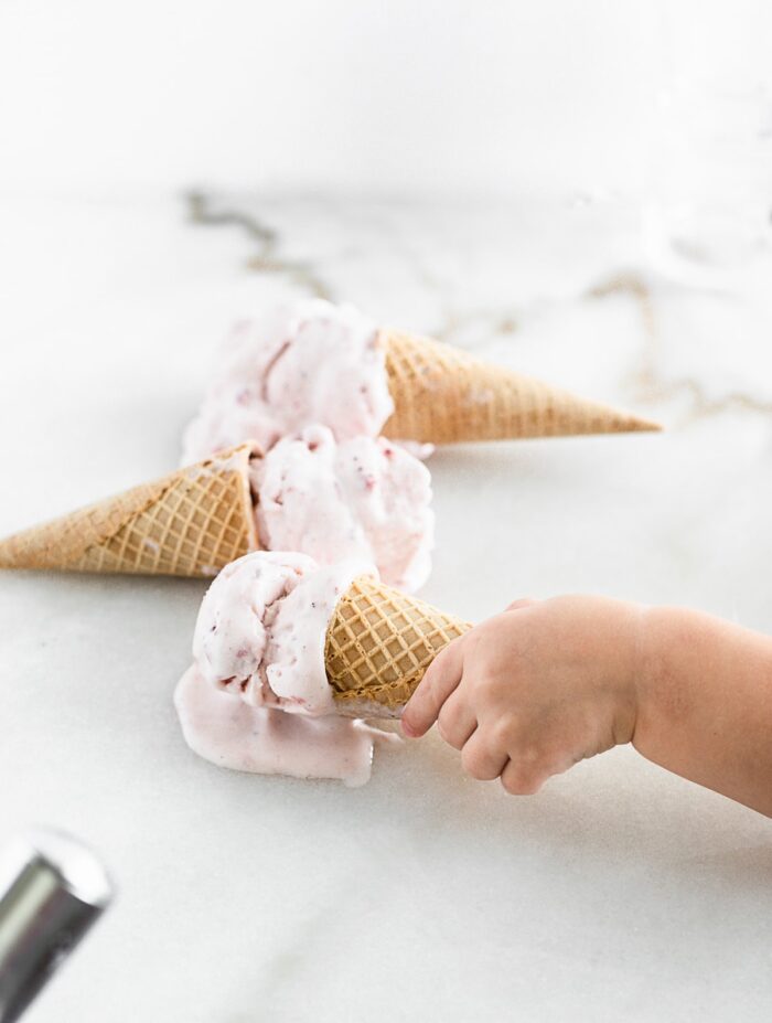 small child's and grabbing a strawberry ice cream cone from a white marble surface with two ice cream cones in the background.