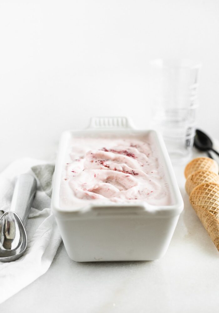 homemade roasted strawberry ice cream in a white loaf pan with an ice cream scoop and ice cream cones on either side.