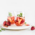 two glasses of rose sangria with peaches and strawberries on a round white marble board.