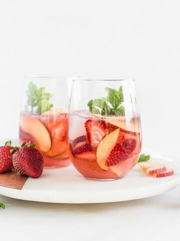 two glasses of rose sangria with peaches and strawberries on a round white marble board.