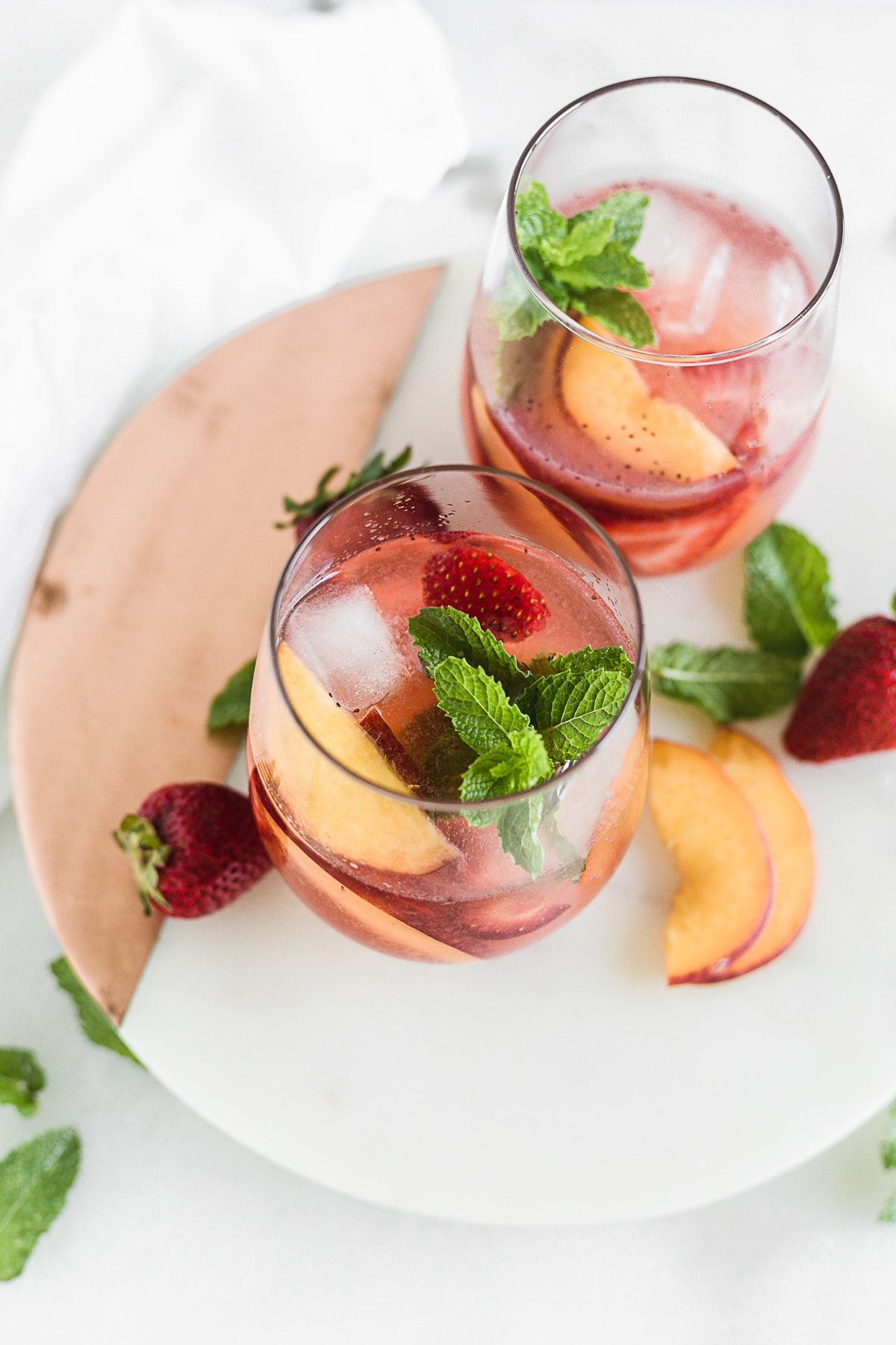 two glasses of rose sangria with peaches and strawberries on a round white marble board surrounded by peach slices and strawberries.