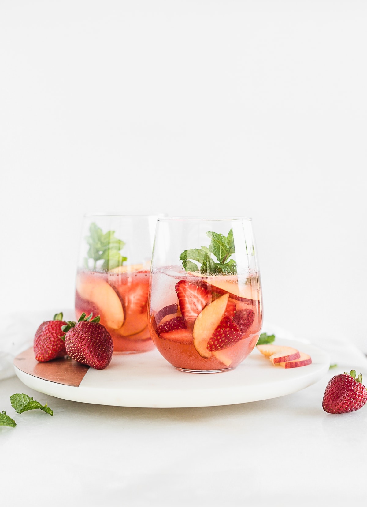 two glasses of rose sangria with peaches and strawberries on a round white marble board.
