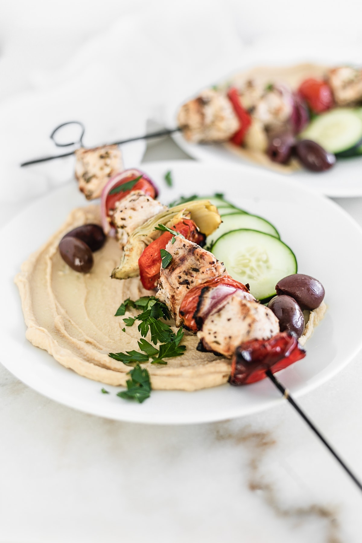 Mediterranean chicken kabobs on plates with hummus, cucumbers, and olives.