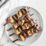 Overhead view of four grilled pineapple Teriyaki pork kabobs on a white plate and a white napkin next to it.