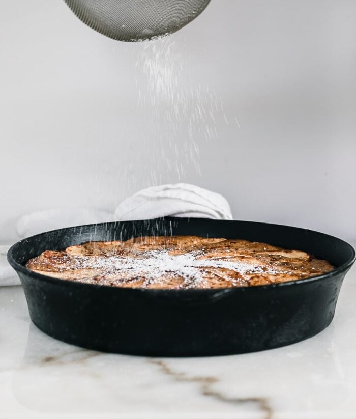 powdered sugar falling onto an apple cake in a cast iron skillet.