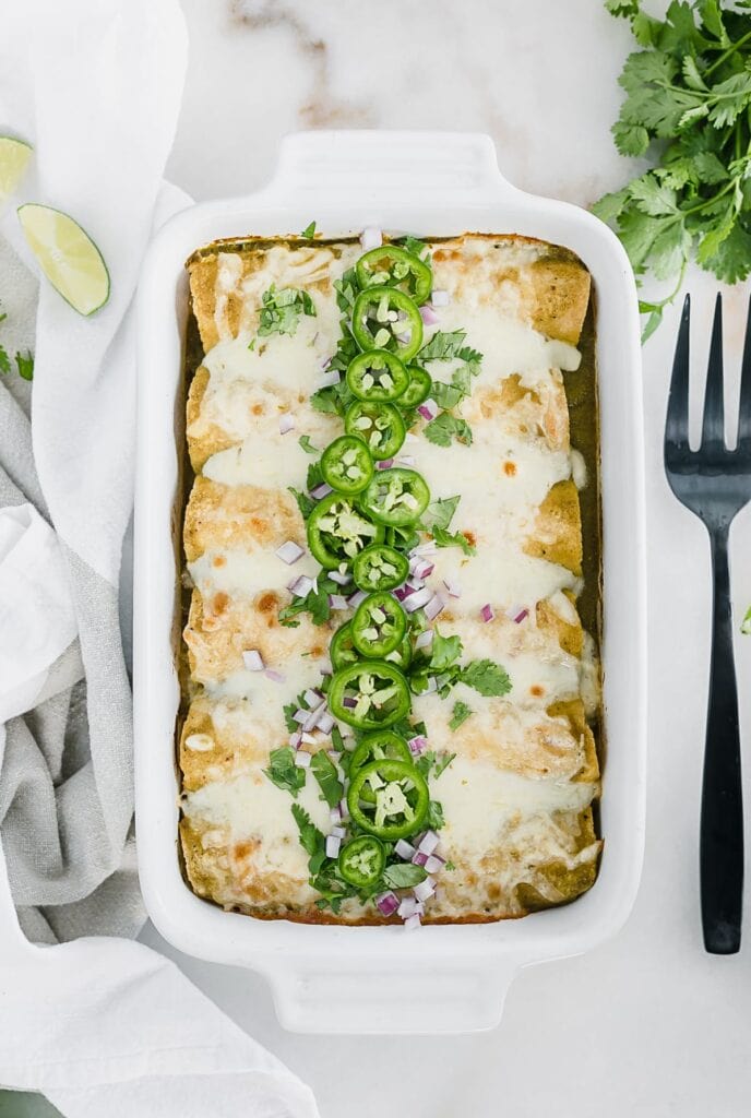 overhead view of Green Chile Chicken Enchiladas in a white baking dish topped with cilantro and jalapenos with a black fork next to the dish.
