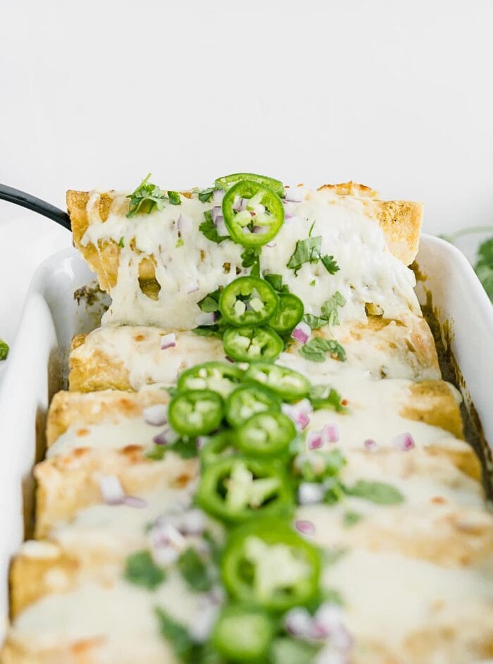 green chile chicken enchiladas topped with cilantro and jalapenos with a black serving spoon pulling one out of the dish.