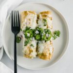 overhead view of two green chile chicken enchiladas on a white plate topped with jalapeno slices and cilantro with a black fork beside them.