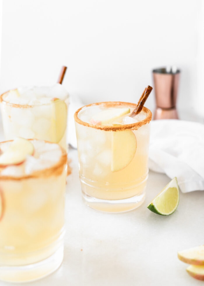 three Apple Cider Margaritas in cinnamon rimmed glasses surrounded by lime slices, a white napkin and a copper jigger.
