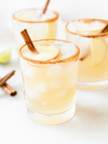 three Apple Cider Margaritas in cinnamon rimmed glasses with apple slices and cinnamon sticks in each.