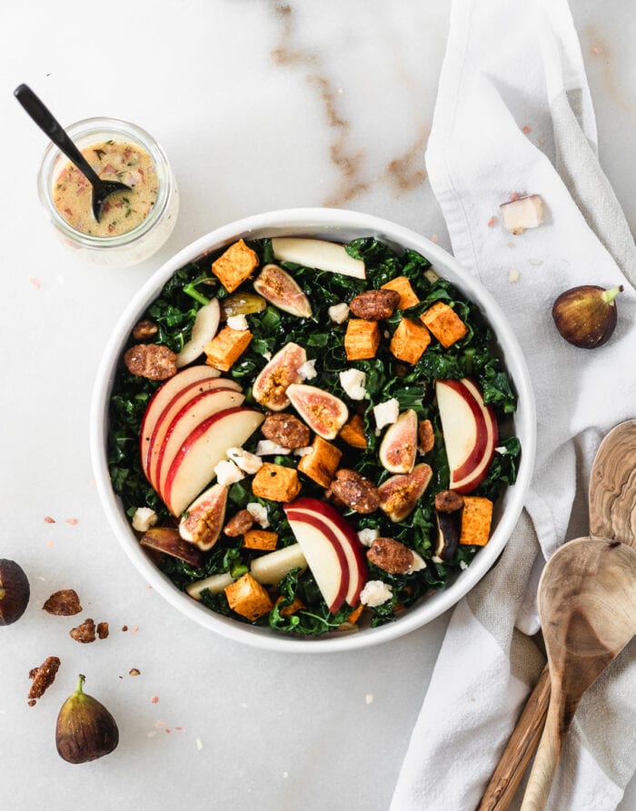 overhead view of warm fall harvest kale salad in a white bowl surrounded by dressing in a jar, wooden spoons, and fresh figs.