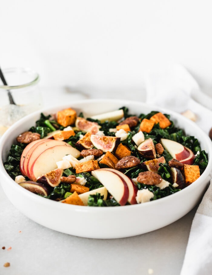 warm fall harvest kale salad with sweet potatoes, apples, candied pecans and figs in a white bowl.
