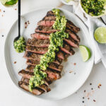 overhead view of sliced grilled flank steak with avocado chimichurri sauce on top on a white plate with a lime wedge and a spoon of sauce.