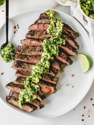 overhead view of sliced grilled flank steak with avocado chimichurri sauce on top on a white plate with a lime wedge and a spoon of sauce.
