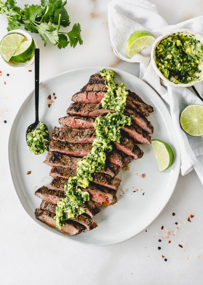 overhead view of sliced grilled flank steak with avocado chimichurri sauce on top on a white plate with a lime wedge and a spoon of sauce.