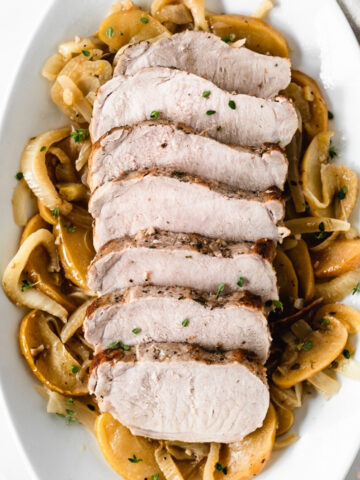 overhead view of sliced pork loin on top of braised apples, onions and fennel on a white platter.