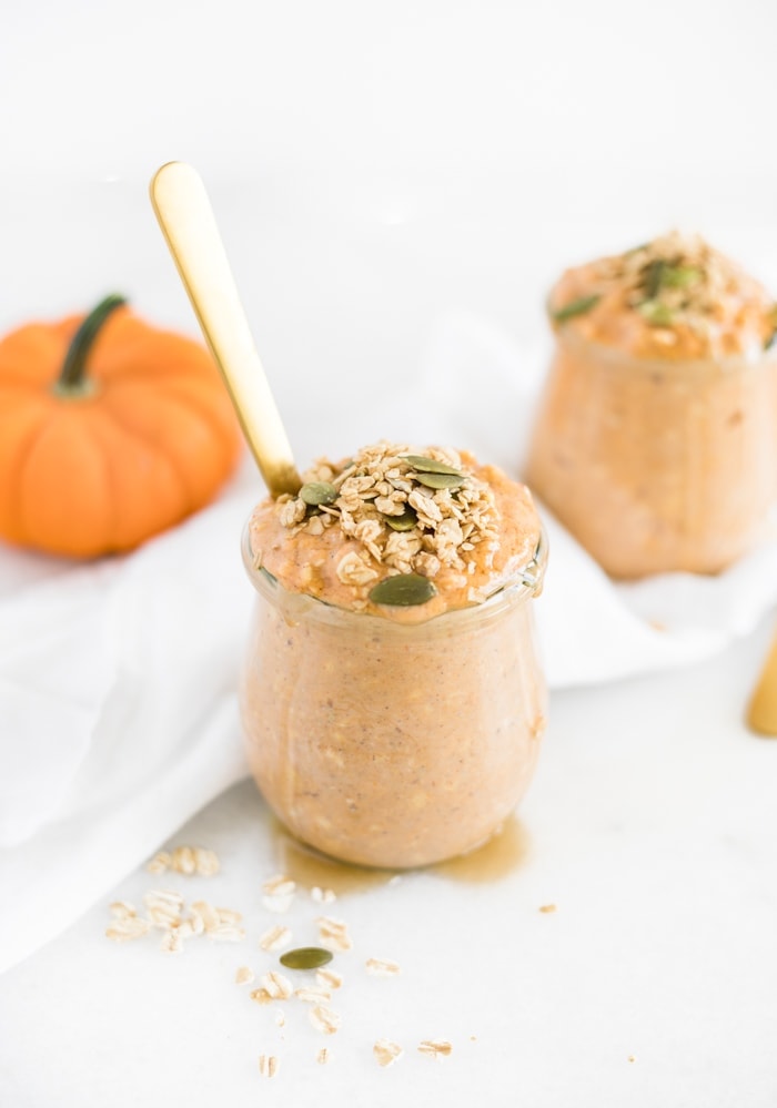 pumpkin spice overnight oats in a glass jar with a gold spoon in it with another jar of oats and a pumpkin behind it.