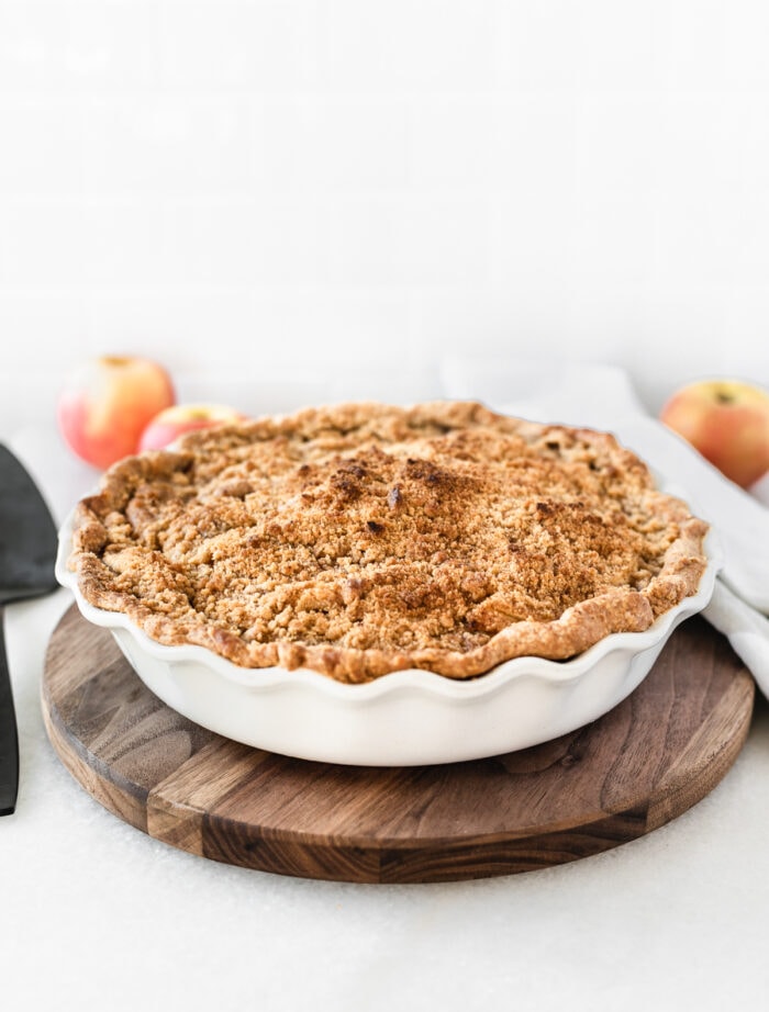 whole wheat apple crumb pie in a white baking dish on a wooden board with apples in the background.