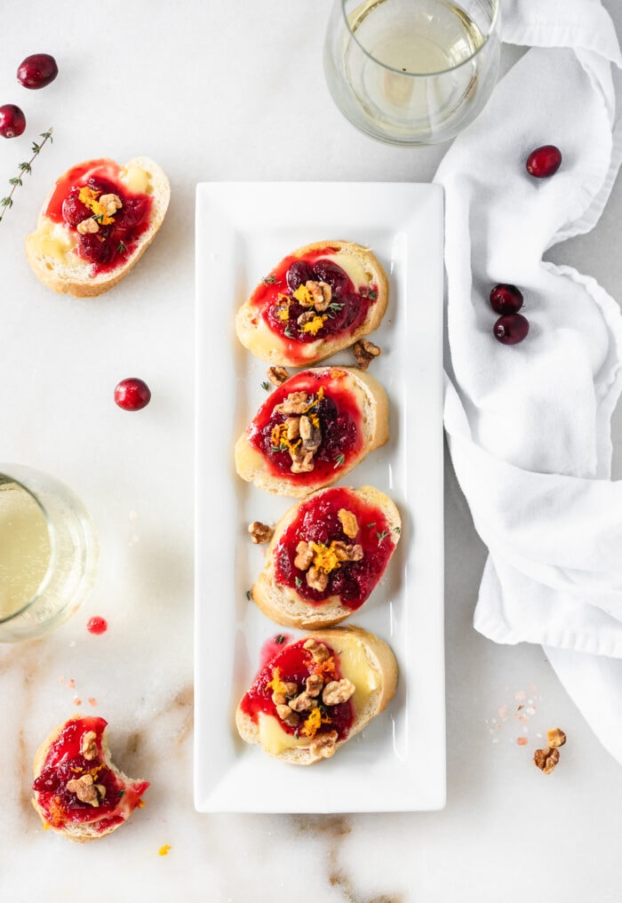 overhead view of cranberry brie crostini lined up on a white serving dish surrounded by wine glasses, walnuts, cranberries, and more crostini.