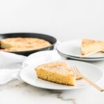 slice of skillet cornbread on a white plate with a gold fork and cornbread skillet behind it.