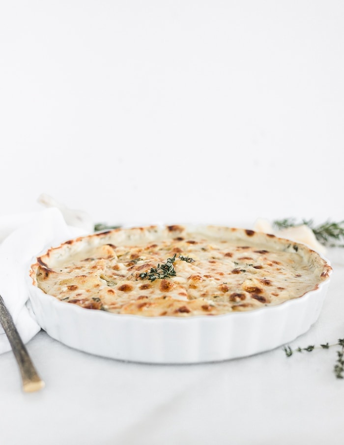 white round baking dish with cheesy scalloped potatoes topped with thyme, surrounded by thyme sprigs, a gold spoon and a white napkin.