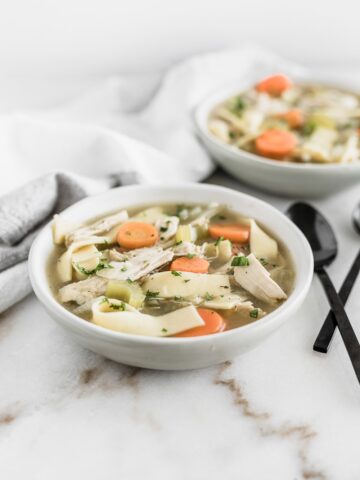 Closeup view of chicken noodle soup in two white bowls with black spoons next to it.