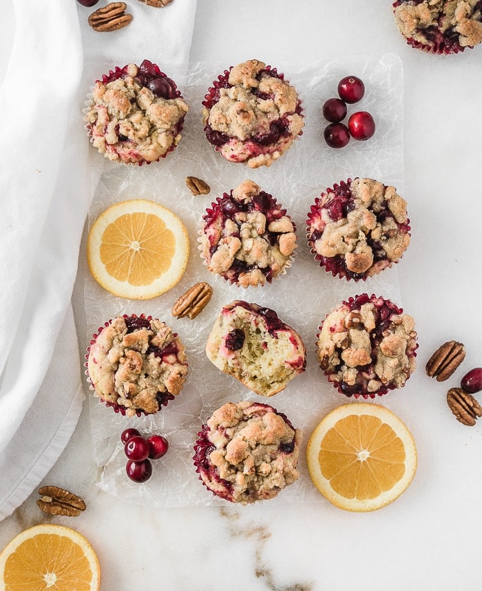 overhead view of cranberry orange crumb muffins on a white background with orange slices, cranberries and pecans.