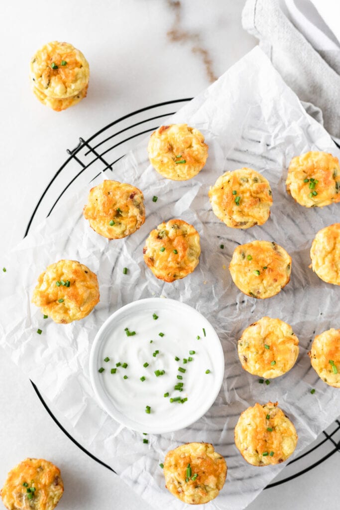 overhead view of mini mashed potato bites on a black wire rack with a bowl of sour cream sprinkled with chives.
