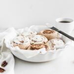 cinnamon rolls with frosting in a round white baking dish lined with parmchment.