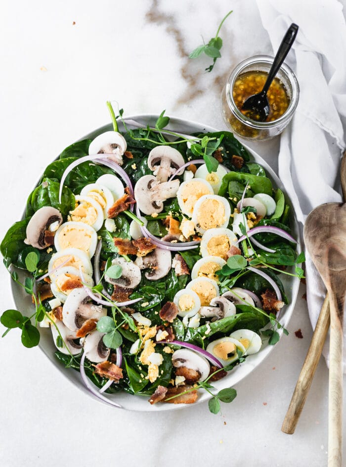 overhead view of mushroom spinach salad with warm sliced boiled eggs, red onion, bacon and warm bacon dressing on a grey plate with wooden spoons beside it..