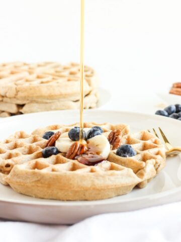 Oatmeal waffle on a white plate with a gold fork topped with pecans, blueberries and sliced bananas getting syrup poured on top.