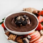 Healthy peanut butter cup cheesecake dip topped with chopped peanut butter cups in a white bowl surrounded by pretzels and fruit.