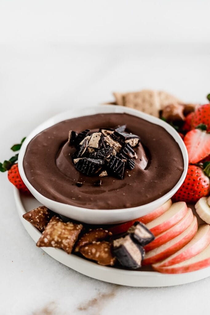 Healthy Peanut Butter Cup Cheesecake Dip topped with chopped peanut butter cups in a white bowl surrounded by pretzels and fruit.