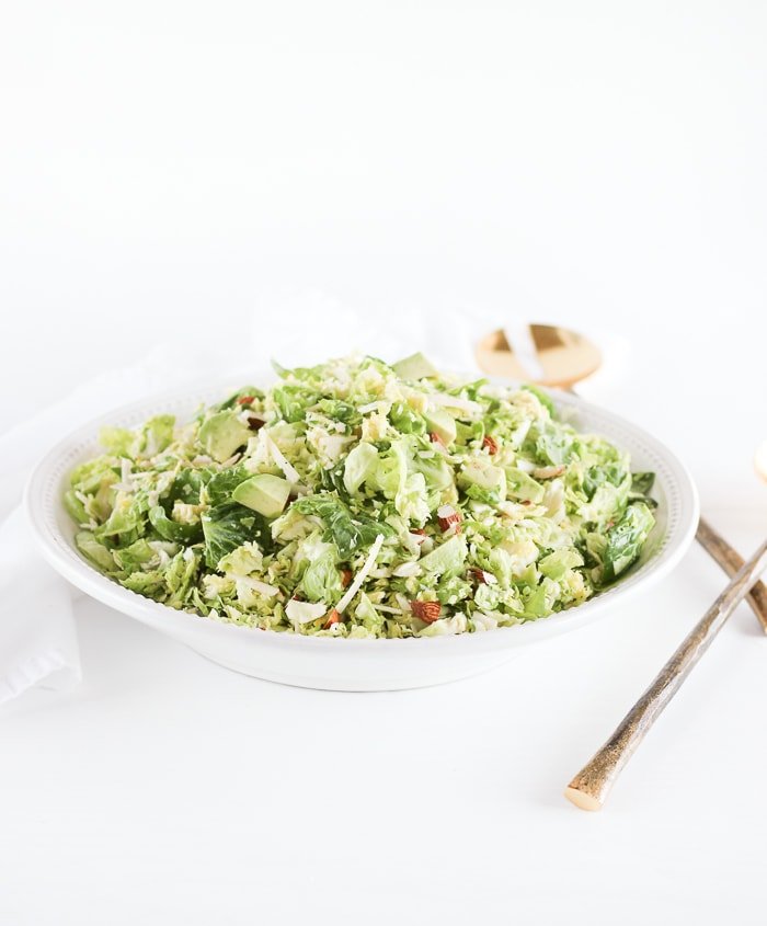 brussels sprouts chopped salad in a white bowl with gold serving spoons in the background.