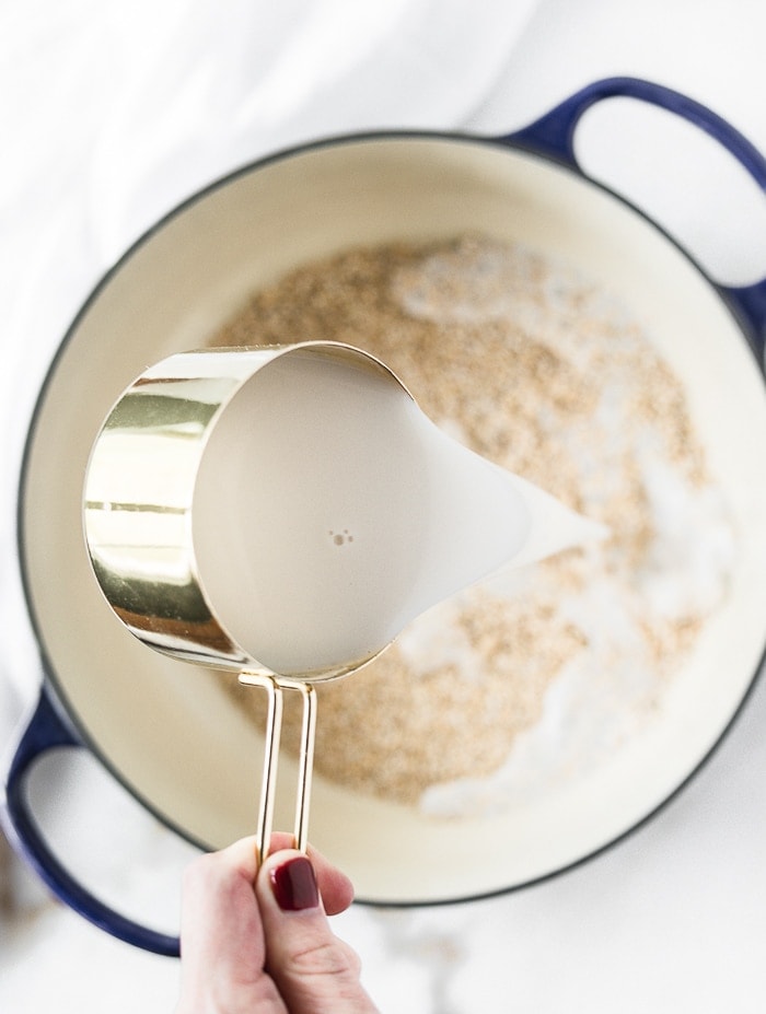 hand pouring a gold measuring cup of milk into a dutch oven with oats.
