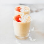 two grapefruit smoothies in glasses each topped with a grapefruit slice and coconut flakes.