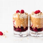 Two glasses with layered PB&J overnight oats with drizzled peanut butter and raspberries on top.