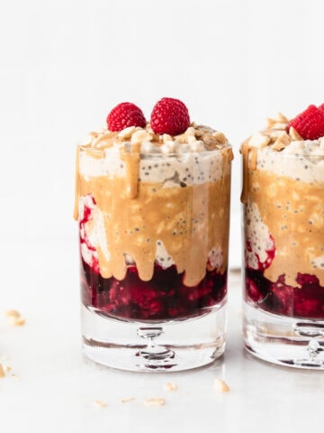 Two glasses with layered PB&J overnight oats with drizzled peanut butter and raspberries on top.