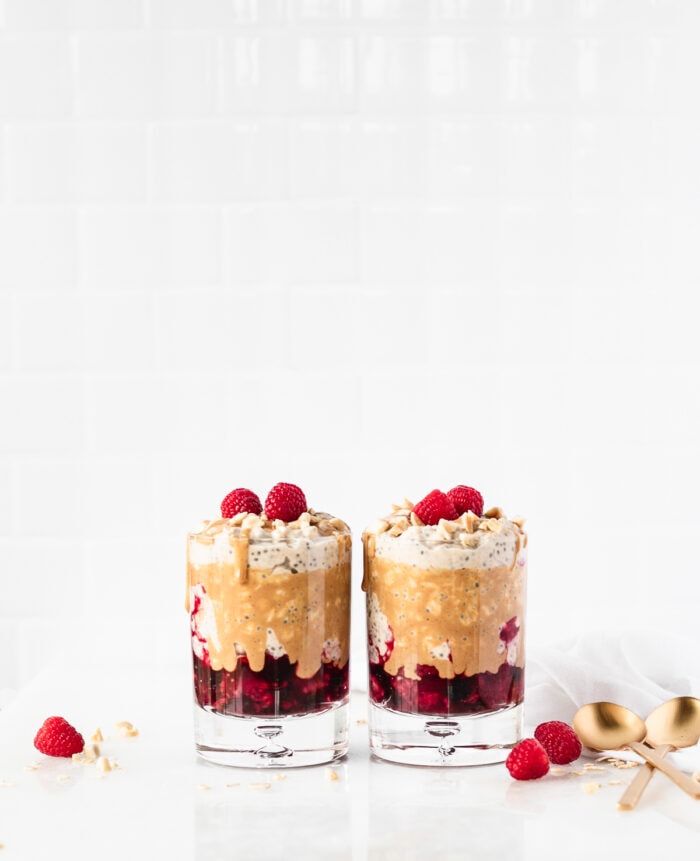 Two glasses with layered PB&J overnight oats with drizzled peanut butter and raspberries on top with gold spoons to the right of them.