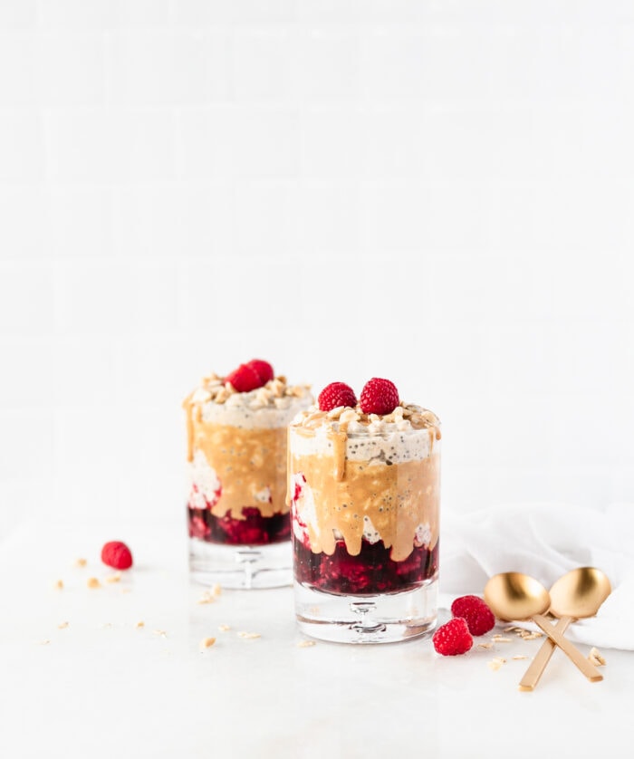 Layered PB&J overnight oats in a glass topped with peanut butter and raspberries with another glass of oat in the background.