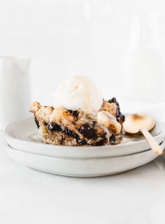 banana bourbon chocolate chip bread pudding with bourbon sauce and ice cream on two stacked plates with a gold spoon next to it.