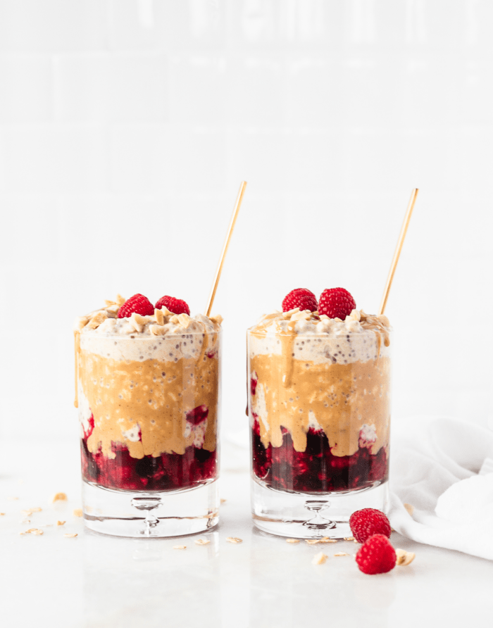 Two glasses with layered PB&J overnight oats with gold spoons in them, with drizzled peanut butter and raspberries on top.