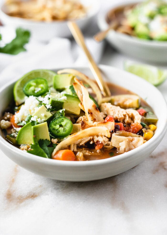 bowl of chicken tortilla soup topped with jalapeno slices, avocado chunks and tortilla strips.