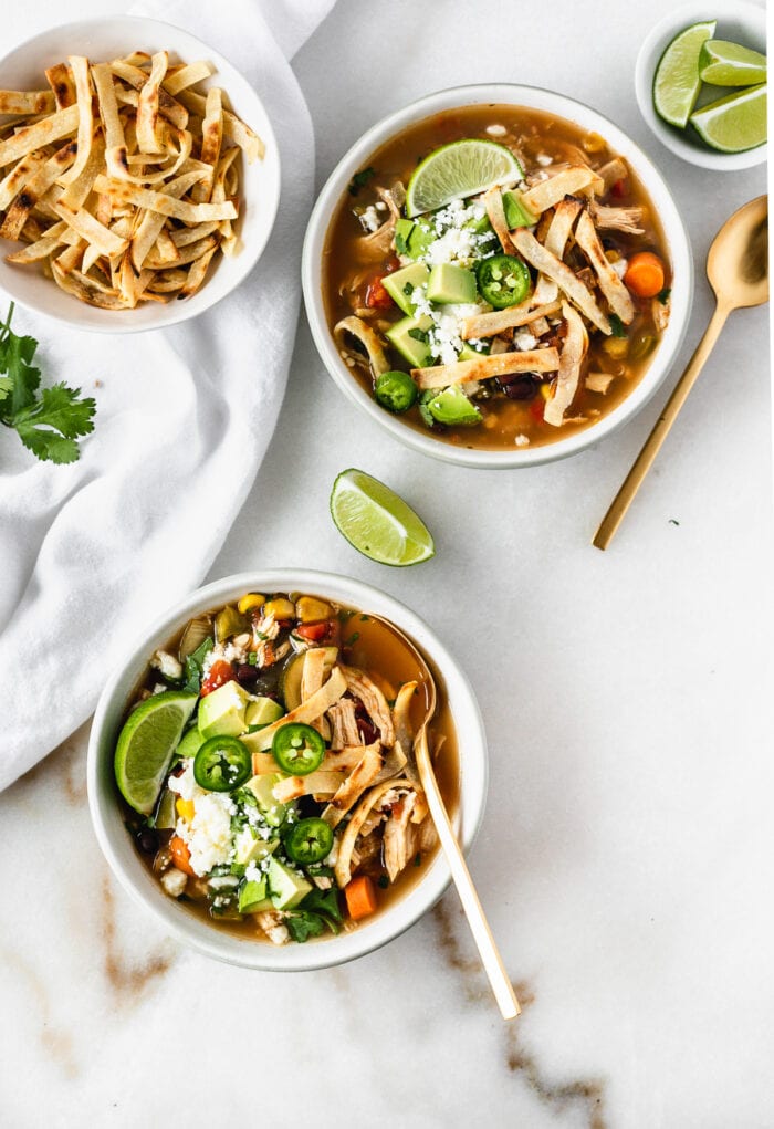 overhead view of two bowls of chicken tortilla soup topped with tortilla strips, jalapeno slices, avocado and limes with gold spoons and a bowl of tortilla strips.