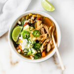 bowl of chicken tortilla soup with avocado, limes, and tortilla strips on top.