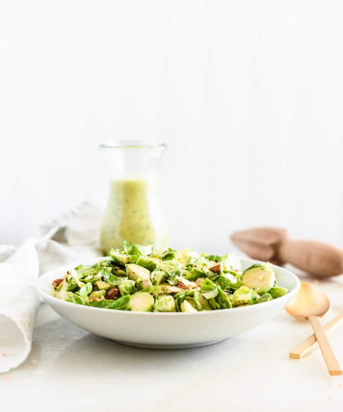 shaved brussels sprouts salad in a white bowl with a glass bottle of citrus shallot dressing behind it.