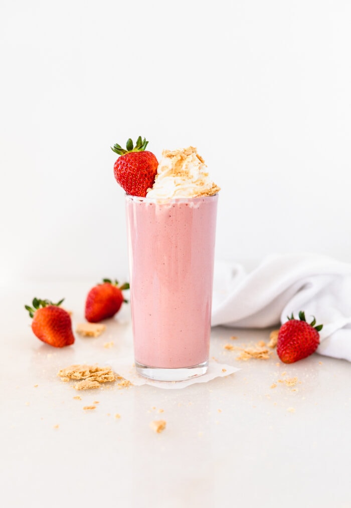 healthy strawberry cheesecake smoothie in a glass topped with whipped cream, graham cracker crumbs and a strawberry.